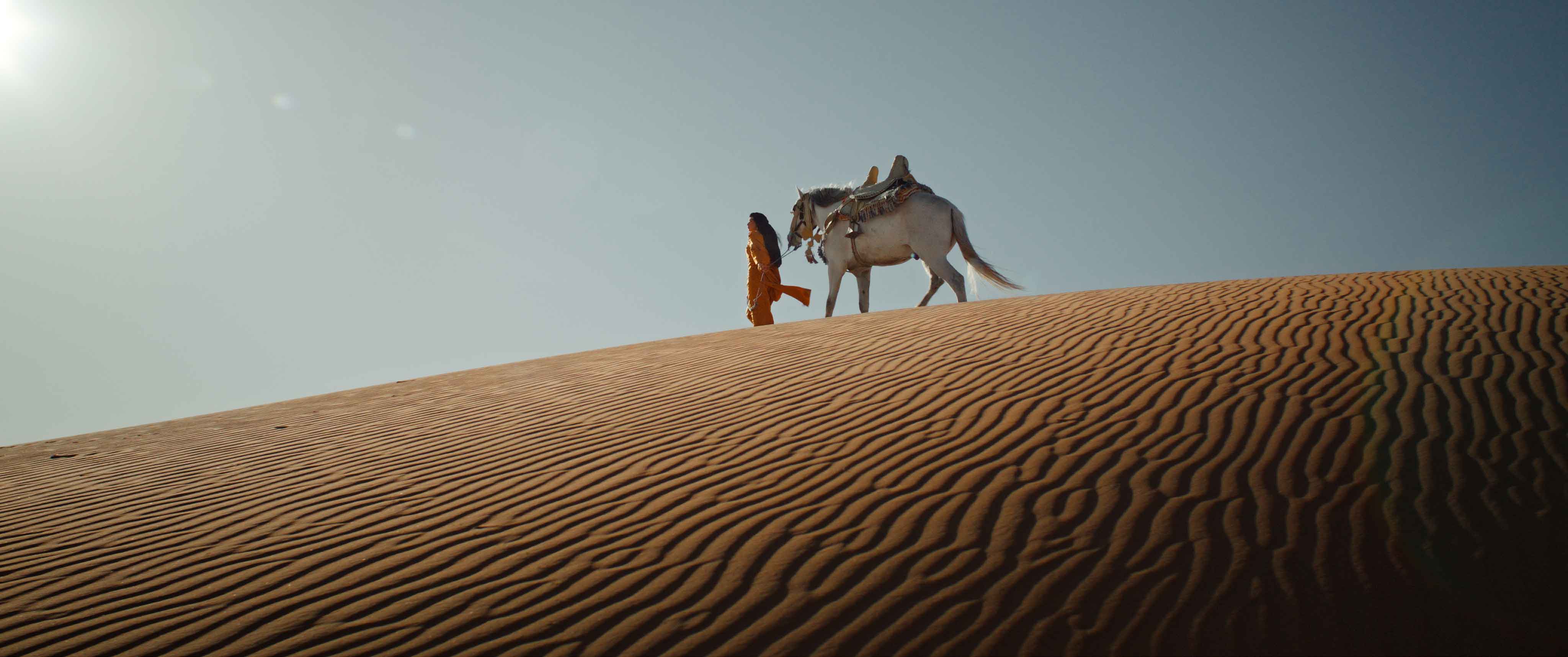 Woman in ochre outfit walking with a white Arabian horse on rippled sand dunes under the desert sun, from Mercedes-Benz MENA Ramadan campaign directed by Farid Malki.